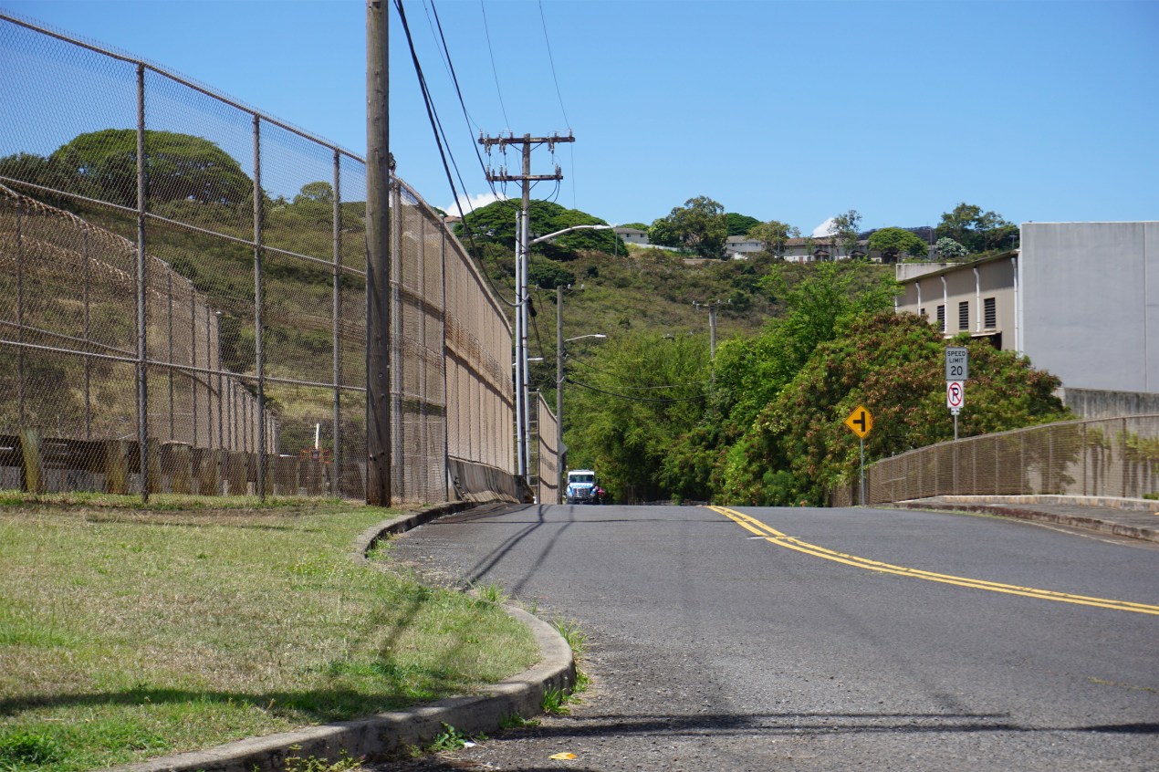 A photo of a road in Honolulu. To the left of the road is a barbed-wire fence, fencing in the Halawa Correctional Facility.