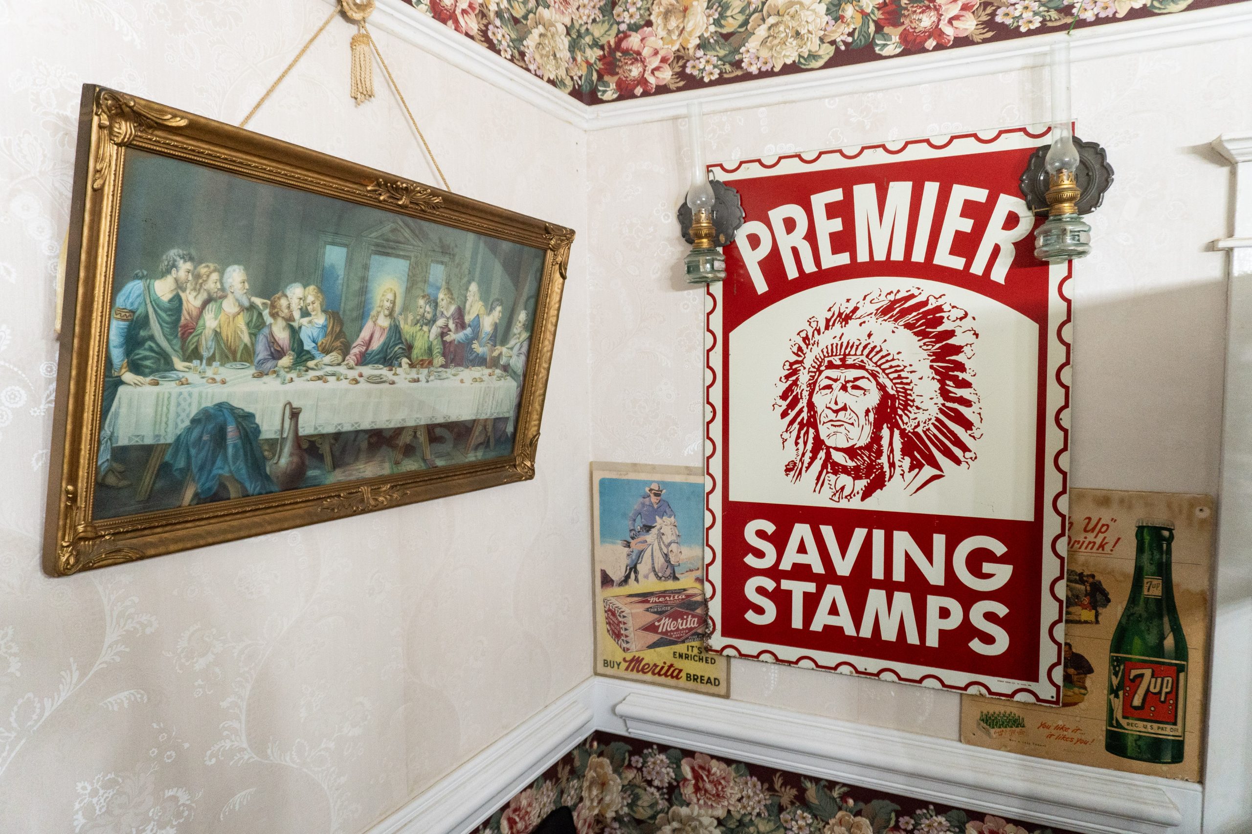 A photo of a wall. On the left is a print of a painting of the Last Supper. On the right are prints of vintage advertisements: Merita bread, a red stamp featuring a Native American man wearing a headdress, and 7Up.