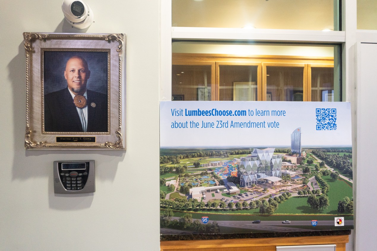 An indoor photo shows a headshot of a John Lowery framed next to a sign that shows a rendering of a casino. Text above it reads, "Visit LumbeesChoose.com to learn more about the June 23rd Amendment vote."
