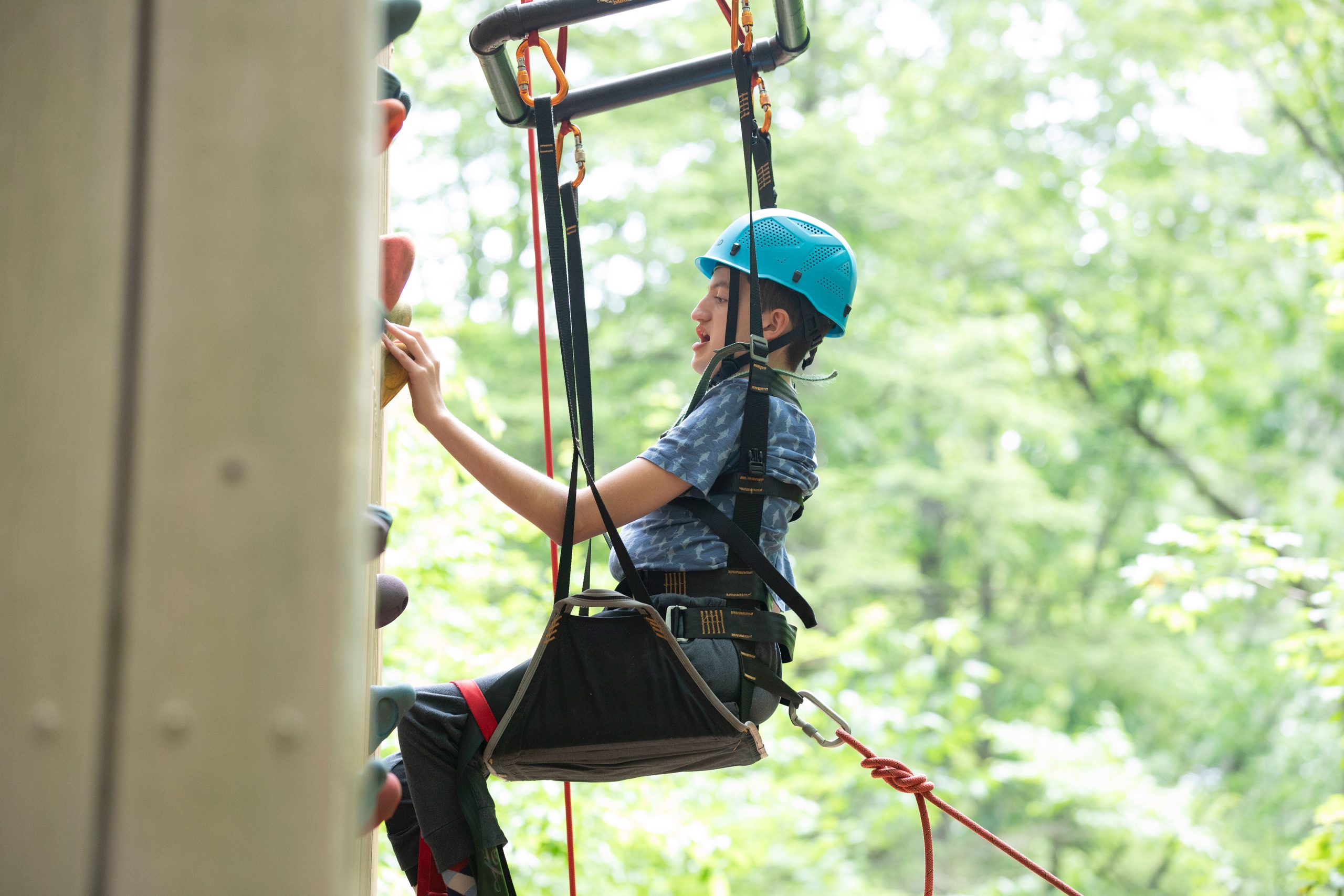 A boy in a blue helmet sits in a harness and holds onto a colorful grip on a rock climbing wall.