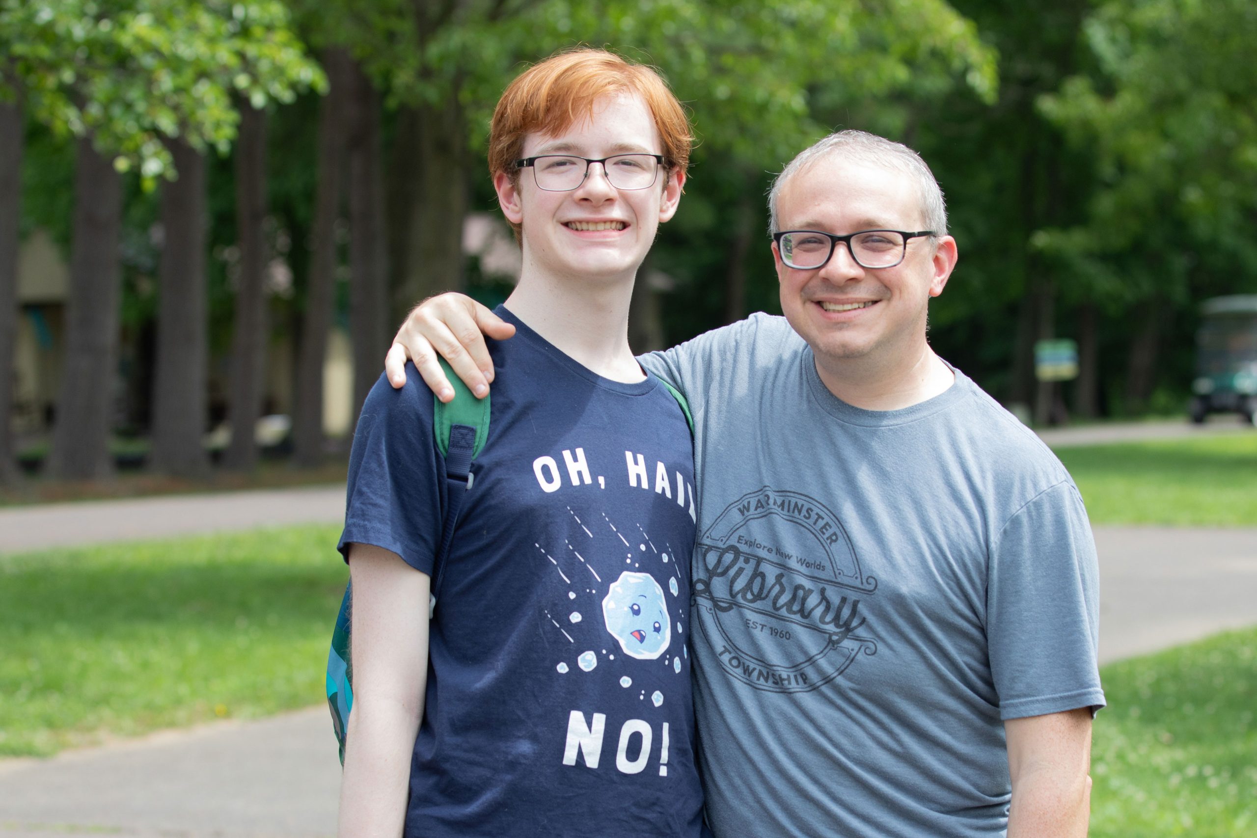 A man stands outside with his arm around the shoulders of his teenage son. Both smile at the camera.